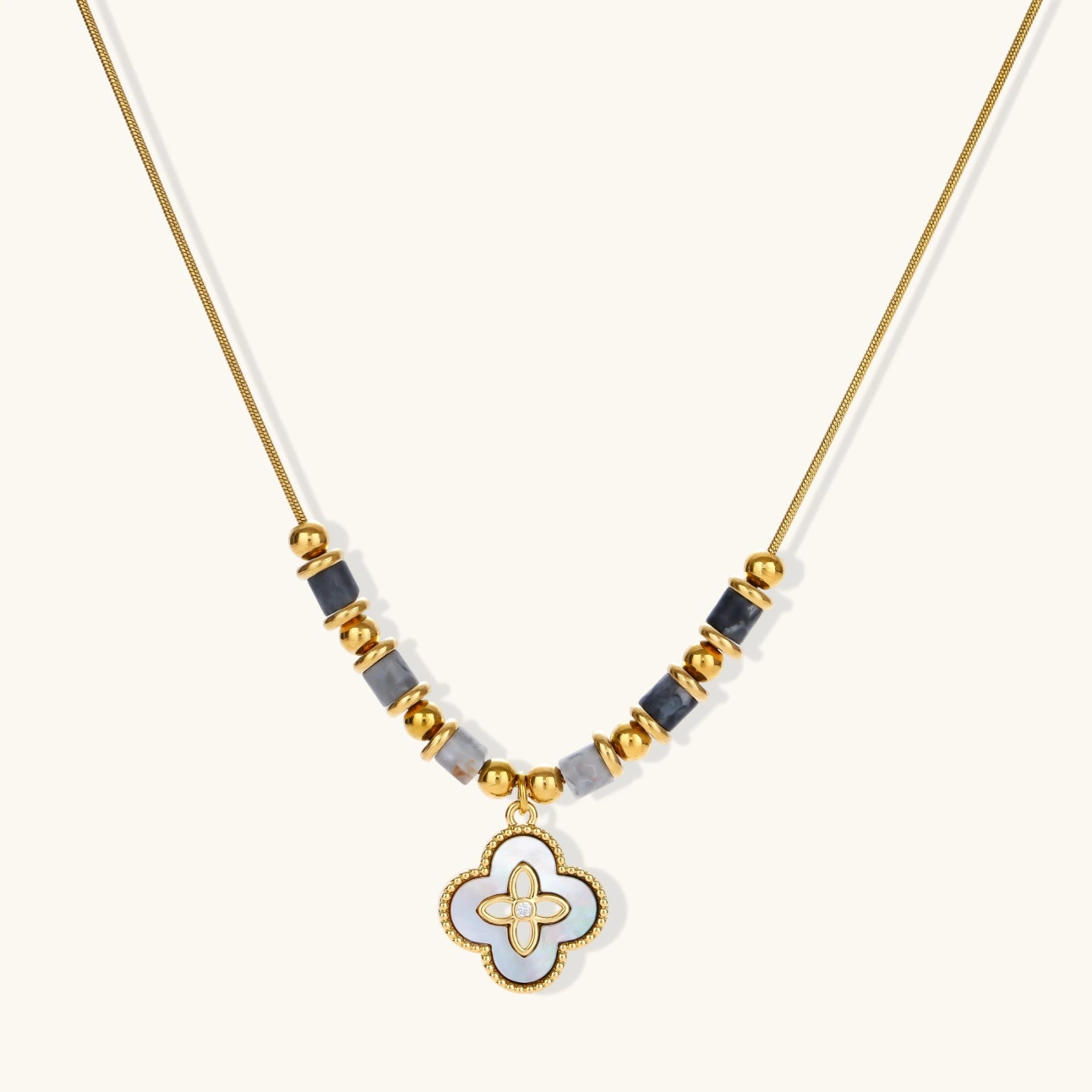 Gold-toned beaded necklace with grey stones and quatrefoil abalone shell pendant on cream background.